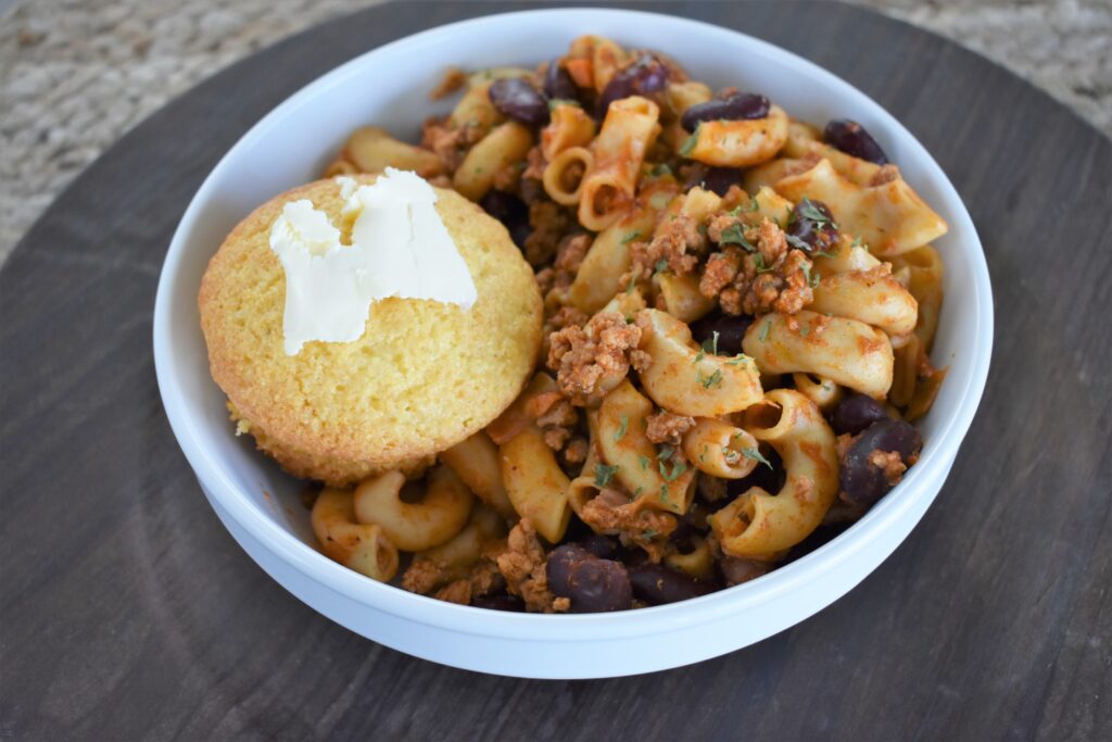 A white bowl holds macaroni pasta with minced meat, kidney beans, and herbs in a red sauce—a hearty meal that can be part of a healthy diet when made with minimal processed foods. Next to it is a cornbread muffin topped with butter, on a dark wooden surface.