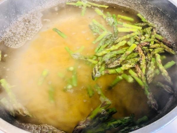 A pot of soup simmers with light brown broth, steam rising from the surface. Fresh green asparagus pieces float on top, reminiscent of a chipotle chicken pasta, surrounded by gentle bubbles and foam along the edges of the saucepan.