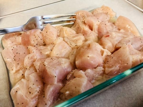 A glass dish filled with raw, cubed chipotle chicken seasoned with black pepper. A metal fork rests on the edge of the dish, partially in the chicken, which is sitting on a light-coloured worktop—perfect for an easy recipe.