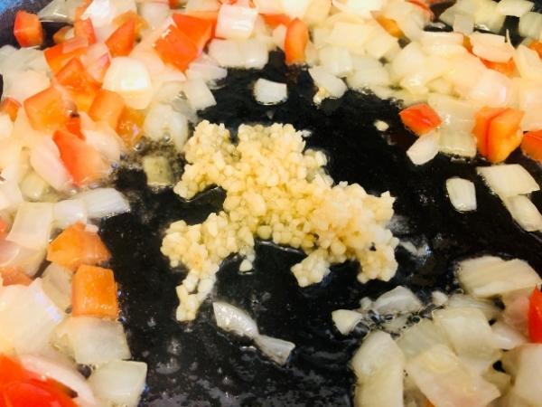 Chopped onions and red peppers sauté in a pan, with a mound of minced garlic in the centre—perfect for starting an easy pasta recipe or adding bold flavour to chipotle chicken pasta. The veg glisten in oil on the pan’s dark surface.