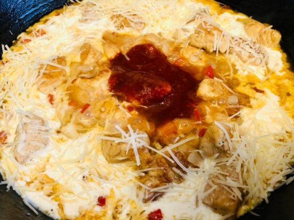 A close-up of an easy Chipotle chicken pasta dish in a pan, featuring cooked chicken, melted cheese, red peppers, and a dollop of tomato sauce. The ingredients blend for a rich, textured appearance with yellow, red, and white tones.