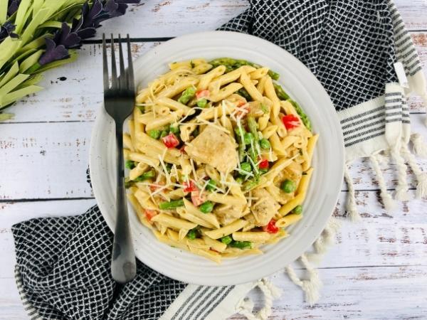 A white plate of easy chicken pasta with penne, grilled chicken, green peas, red peppers, and grated cheese sits on a black-and-white chequered serviette. A black fork rests on the plate atop a rustic white wooden surface with green leaves in the corner.