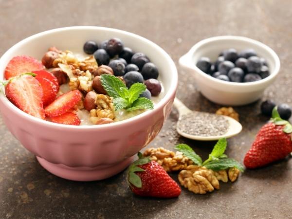 A pink bowl filled with porridge, topped with sliced strawberries, blueberries, walnuts, hazelnuts, and mint—a perfect back to school breakfast. Nearby are fresh strawberries, a wooden spoon of chia seeds, blueberries in a bowl, and scattered walnuts and mint leaves.