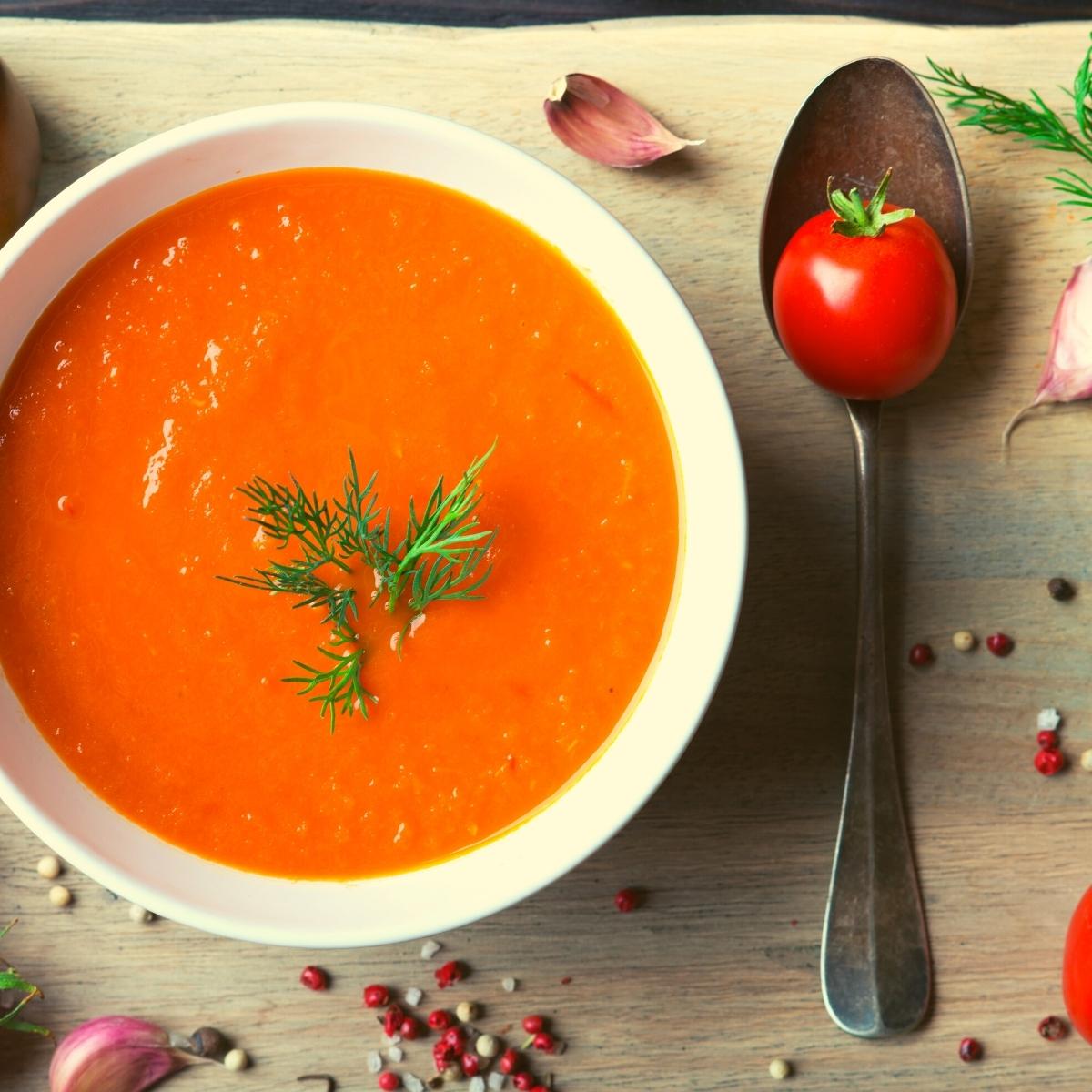 A bowl of bright orange tomato soup garnished with fresh dill sits on a wooden surface. Surrounded by garlic, peppercorns, and a cherry tomato, it invites you to savour the flavours of home gardening and discover delicious tomato tips.