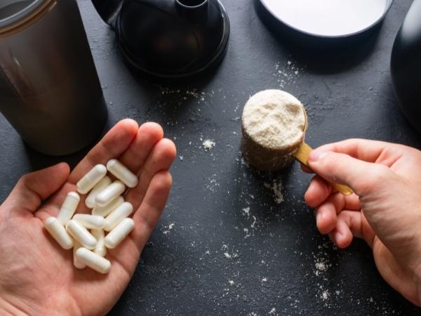 A hand holds several white supplement capsules, while the other grasps a scoop of protein powder. The scene, set against a dark surface with scattered powder and shaker bottles, evokes superfood benefits and the latest superfood trends in fitness nutrition.