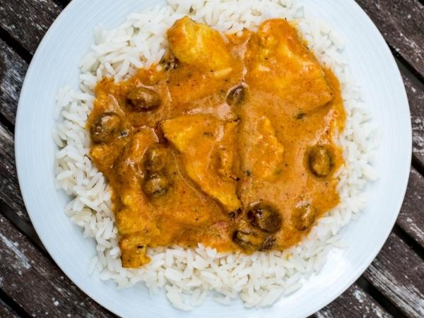 A white plate on a wooden table holds white rice topped with a creamy orange curry. The curry, perfect for beating fatigue when cooking, has chunks of fish and small mushrooms in its thick, flavourful sauce spread over the rice.