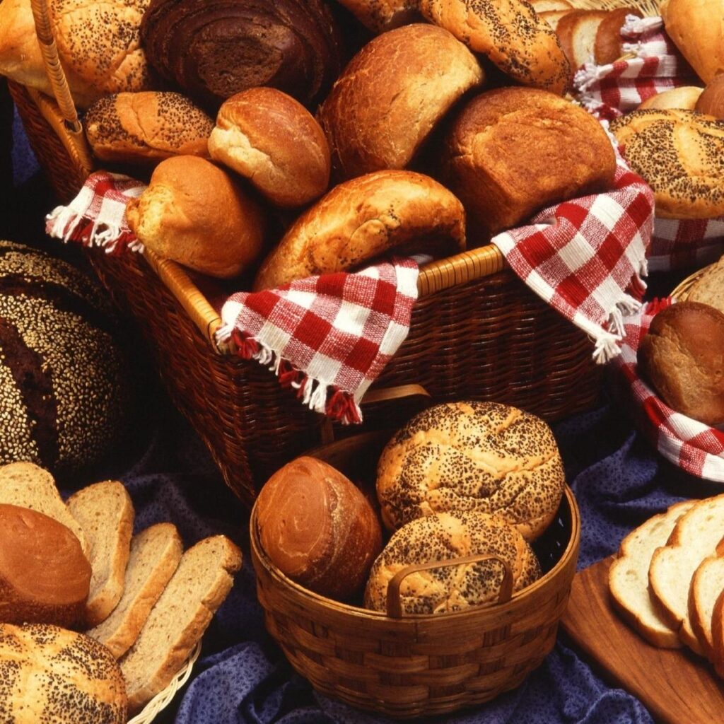 A wicker basket lined with red and white checked cloth is filled with assorted bread rolls, each one baked to perfection. Surrounding it, more baskets display breads that would brighten any kitchen, from sliced loaves to seeded buns on a dark blue cloth.