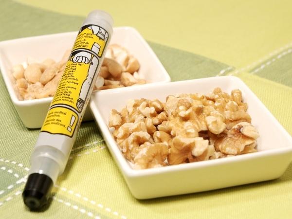 A yellow adrenaline auto-injector pen rests beside two small dishes—one with shelled walnuts, the other with peanuts—on a green and yellow tablecloth, highlighting the importance of managing food allergies in everyday settings.