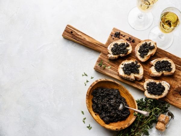A wooden board holds five slices of bread topped with black caviar, an exotic food, next to a bowl filled with more caviar and a spoon. Two glasses of white wine and sprigs of fresh herbs are nearby, all set on a light, textured surface.