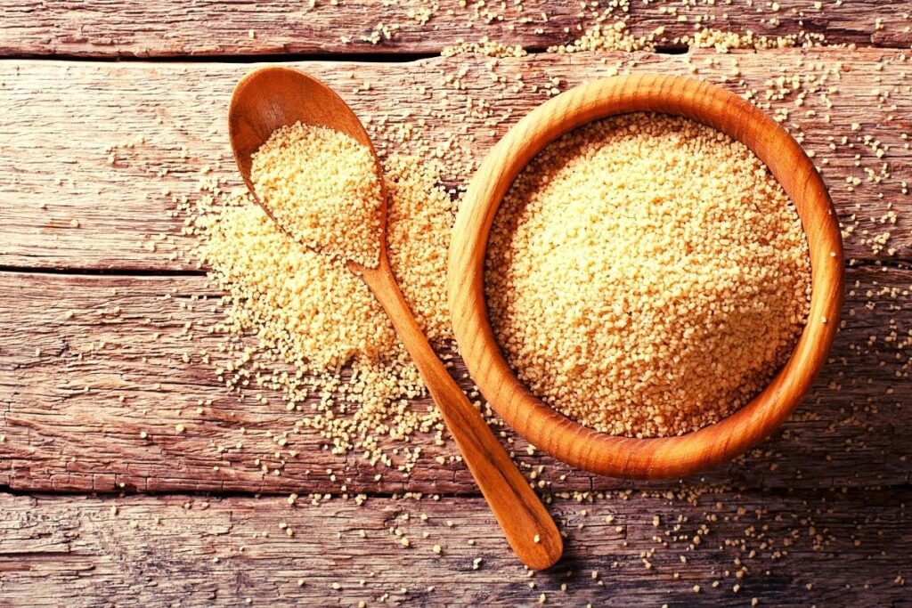 A wooden bowl filled with uncooked couscous sits on a rustic wooden surface. Next to the bowl, a wooden spoon holds more couscous, suggesting fresh ingredients ready for a delicious cold couscous salad in a natural, earthy presentation.