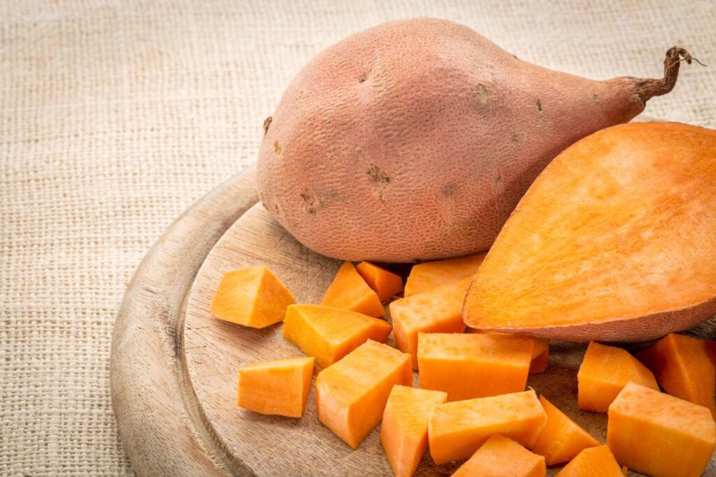 A whole sweet potato, a halved sweet potato showing its bright orange flesh, and several cubed sweet potatoes are arranged on a wooden chopping board atop a textured beige fabric surface, ready for cooking.