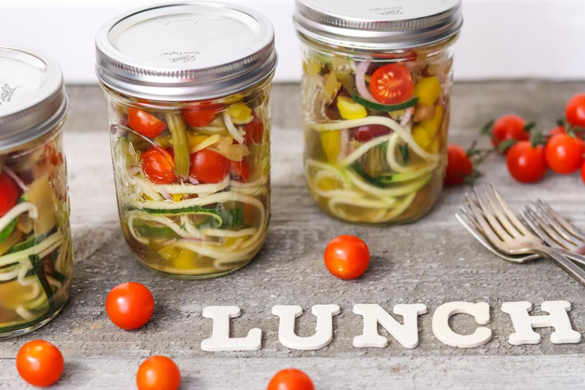 Three Kilner jars filled with colourful salad—cherry tomatoes, spiralised veg, and greens—sit on a wooden surface. Loose tomatoes, forks, and white letters spelling “LUNCH” complete this fresh idea for healthy recipes.