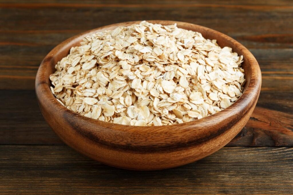 A wooden bowl filled with the best oats sits on a dark wooden surface. The light tan, flat oats have a rough texture, making them perfect for breakfast. The rustic wood grain complements their natural, unprocessed appearance.