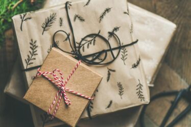 Three presents wrapped in brown and beige paper with botanical patterns and string bows are stacked on a wooden surface, perfect as a gift for foodies. Scissors and evergreen branches nearby suggest a cosy, kitchen-inspired gift-wrapping scene.