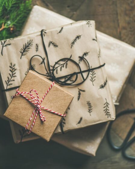 Three presents wrapped in brown and beige paper with botanical patterns and string bows are stacked on a wooden surface, perfect as a gift for foodies. Scissors and evergreen branches nearby suggest a cosy, kitchen-inspired gift-wrapping scene.