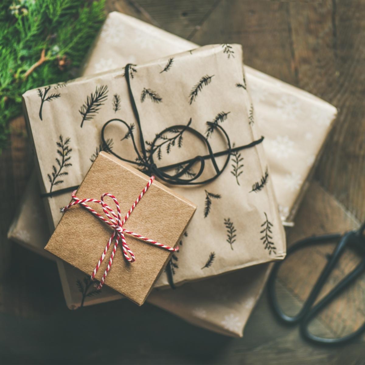 Three presents wrapped in brown and beige paper with botanical patterns and string bows are stacked on a wooden surface, perfect as a gift for foodies. Scissors and evergreen branches nearby suggest a cosy, kitchen-inspired gift-wrapping scene.