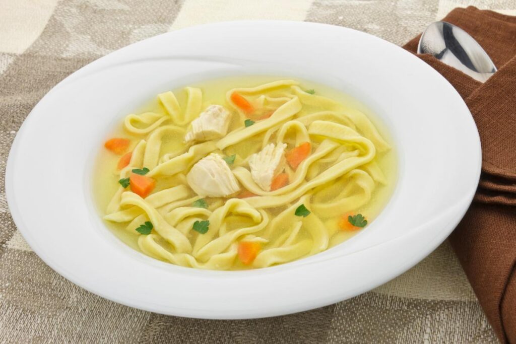 A white bowl of homemade chicken noodle soup with wide egg noodles, chunks of chicken, diced carrots, and parsley in a clear broth sits on a beige chequered tablecloth next to a brown serviette and a spoon.