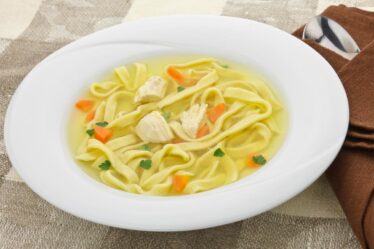 A white bowl of homemade chicken noodle soup with wide egg noodles, chunks of chicken, diced carrots, and parsley in a clear broth sits on a beige chequered tablecloth next to a brown serviette and a spoon.