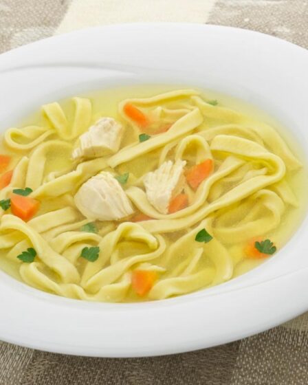 A white bowl of homemade chicken noodle soup with wide egg noodles, chunks of chicken, diced carrots, and parsley in a clear broth sits on a beige chequered tablecloth next to a brown serviette and a spoon.