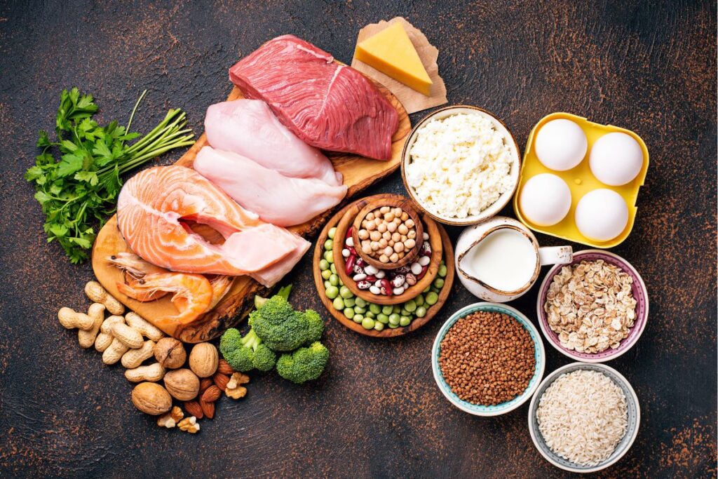 A selection of high-protein food displayed on a dark surface, featuring raw salmon, chicken, beef, eggs, cheese, milk, cottage cheese, beans, nuts, oats, rice, lentils, broccoli and fresh parsley neatly arranged with bowls and wooden boards.