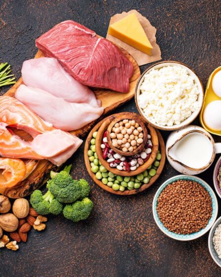 A selection of high-protein food displayed on a dark surface, featuring raw salmon, chicken, beef, eggs, cheese, milk, cottage cheese, beans, nuts, oats, rice, lentils, broccoli and fresh parsley neatly arranged with bowls and wooden boards.