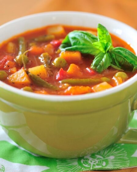 A green ceramic cup filled with chunky vegetable soup, featuring visible green beans, carrots, potatoes, and tomatoes, garnished with basil. For soup tips like thickening soup if it’s too runny, the cup sits on a folded green and white serviette on a wooden tray.
