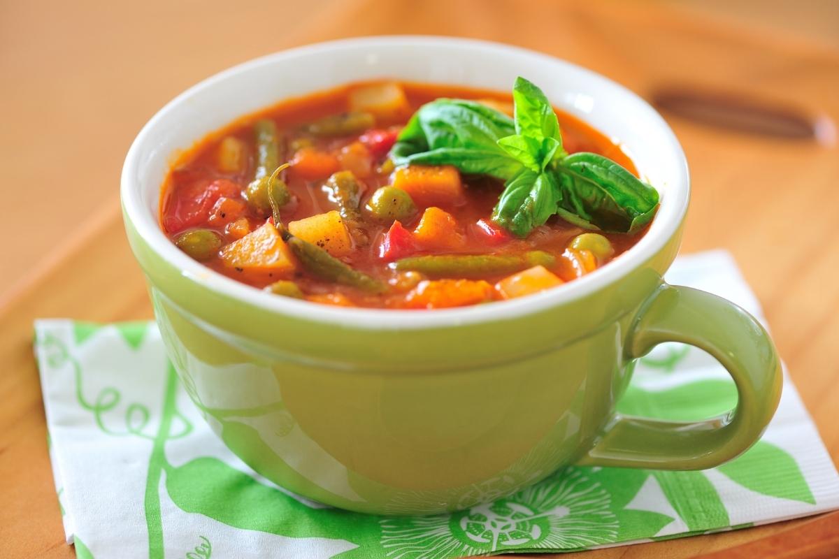 A green ceramic cup filled with chunky vegetable soup, featuring visible green beans, carrots, potatoes, and tomatoes, garnished with basil. For soup tips like thickening soup if it’s too runny, the cup sits on a folded green and white serviette on a wooden tray.