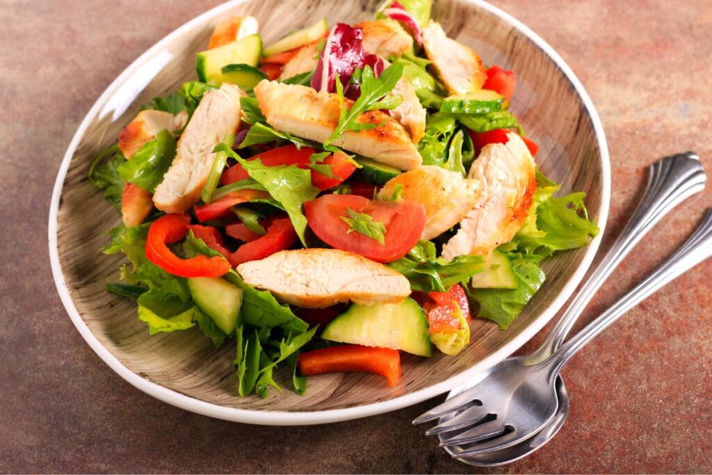 A plate of mixed salad with grilled chicken strips, fresh veg, and rocket sits on a wooden-patterned plate. The colourful dish pairs perfectly with easy salad dressings for a quick, flavourful meal beside a fork and spoon on a brown surface.