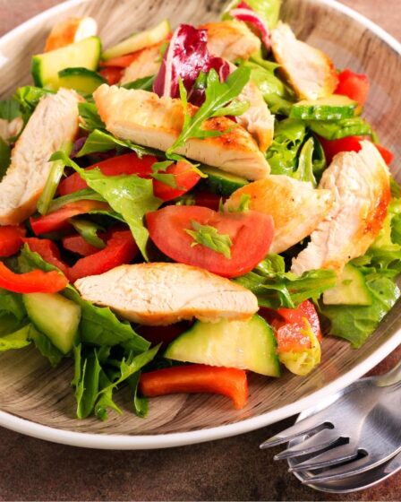 A plate of mixed salad with grilled chicken strips, fresh veg, and rocket sits on a wooden-patterned plate. The colourful dish pairs perfectly with easy salad dressings for a quick, flavourful meal beside a fork and spoon on a brown surface.