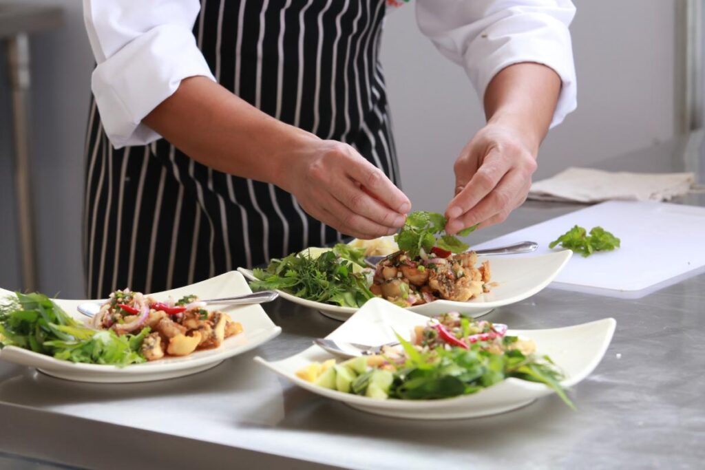 A chef in a striped apron and white sleeves carefully places fresh herbs on plated salads inspired by Emirati cuisine in a commercial kitchen. Three dishes, garnished with greens and vegetables, sit on a stainless steel counter beside more herbs.