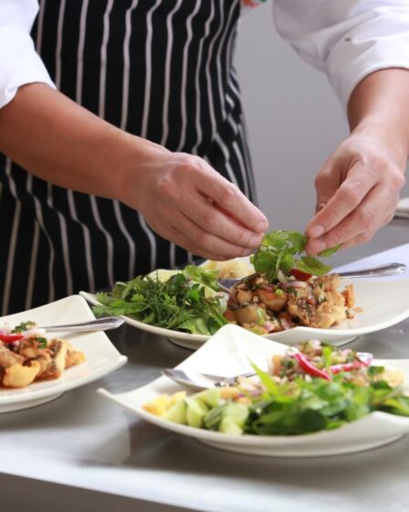A chef in a striped apron and white sleeves carefully places fresh herbs on plated salads inspired by Emirati cuisine in a commercial kitchen. Three dishes, garnished with greens and vegetables, sit on a stainless steel counter beside more herbs.