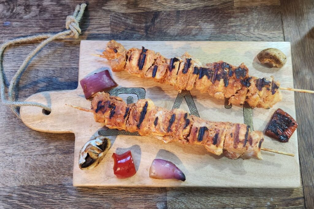 Two grilled chicken skewers with visible grill marks rest on a wooden chopping board, surrounded by assorted grilled vegetables. Perfect as children's meals or for picky eater meals, the rustic table and warm lighting make this dish inviting for all.