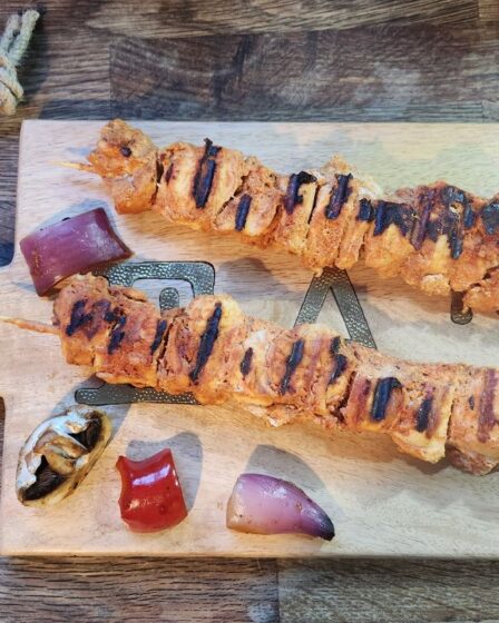 Two grilled chicken skewers with visible grill marks rest on a wooden chopping board, surrounded by assorted grilled vegetables. Perfect as children's meals or for picky eater meals, the rustic table and warm lighting make this dish inviting for all.