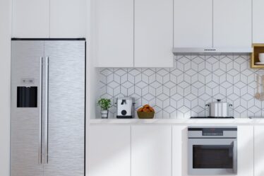 Modern white kitchen maximises cooking space with a stainless steel fridge, built-in oven, hob, and hood. A coffee maker and fruit bowl sit on the worktop. Geometric tile splashback complements the light wood herringbone floor.