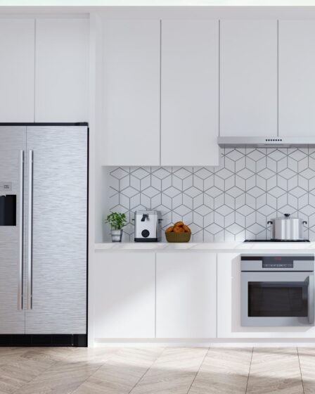 Modern white kitchen maximises cooking space with a stainless steel fridge, built-in oven, hob, and hood. A coffee maker and fruit bowl sit on the worktop. Geometric tile splashback complements the light wood herringbone floor.