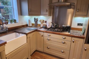 Modern kitchen with cream cabinets, wooden worktops, and stainless steel appliances—ideal for packing kitchen essentials during a house move. A butler sink sits under a window with potted plants, while wooden flooring completes the cosy look.