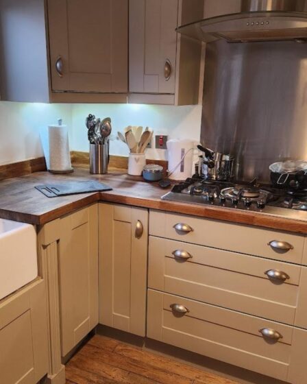 Modern kitchen with cream cabinets, wooden worktops, and stainless steel appliances—ideal for packing kitchen essentials during a house move. A butler sink sits under a window with potted plants, while wooden flooring completes the cosy look.