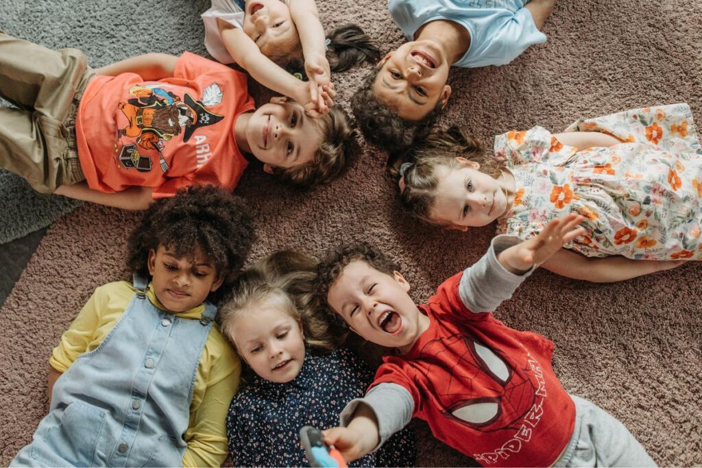 Six young children lie in a circle on a carpet, heads together and smiling happily. Their colourful outfits and joyful expressions reflect the diversity that fosters resilience and supports child development through play and connection.