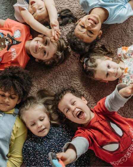 Six young children lie in a circle on a carpet, heads together and smiling happily. Their colourful outfits and joyful expressions reflect the diversity that fosters resilience and supports child development through play and connection.
