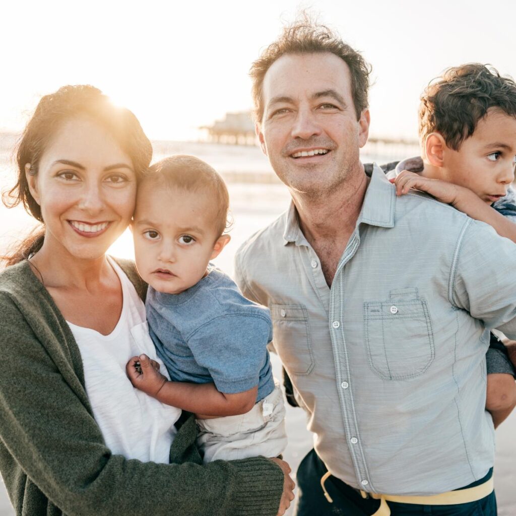 A smiling woman and man stand close together outdoors, each holding a young child. The sunlit background suggests a beach setting, perfect for summer family fun. Everyone appears happy and relaxed, enjoying cherished family activities together.