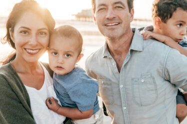 A smiling woman and man stand close together outdoors, each holding a young child. The sunlit background suggests a beach setting, perfect for summer family fun. Everyone appears happy and relaxed, enjoying cherished family activities together.