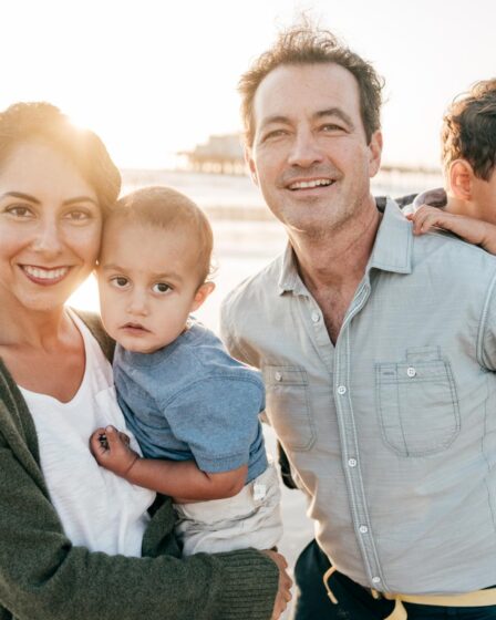 A smiling woman and man stand close together outdoors, each holding a young child. The sunlit background suggests a beach setting, perfect for summer family fun. Everyone appears happy and relaxed, enjoying cherished family activities together.