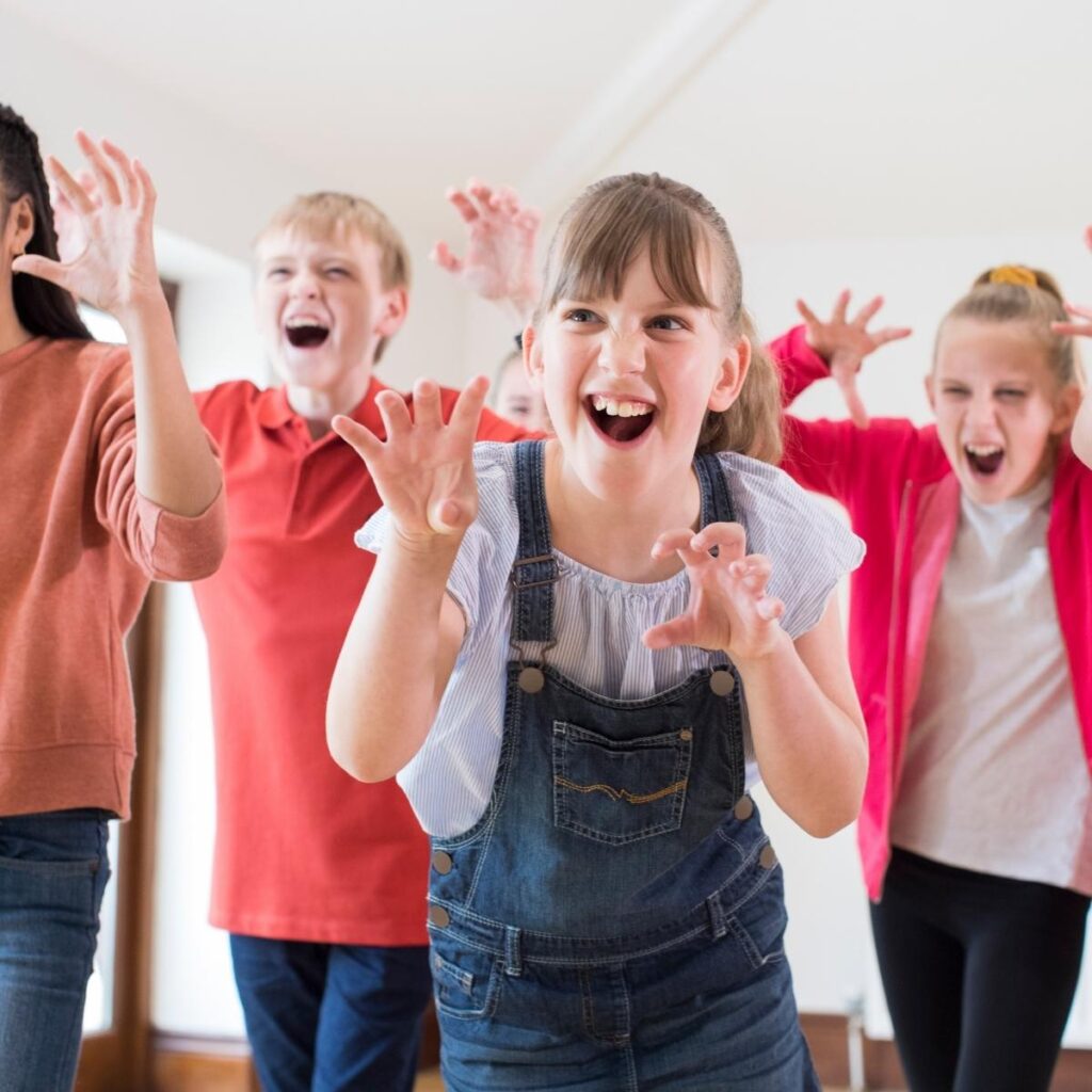 Four children indoors laugh and pretend to be scary monsters, raising their hands with clawed fingers and playful expressions. This lively scene highlights how sensory activities can spark creativity and fun, catering to various children's sensory needs.