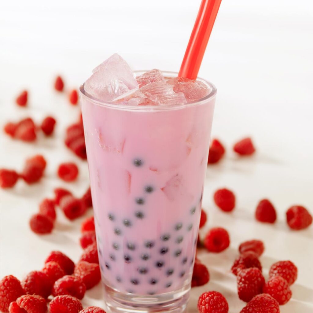 A tall glass of pink bubble tea with black tapioca pearls and ice cubes, served with a red straw. Fresh raspberries are scattered around the glass on a white surface, inspiring you to make bubble tea at home. The softly blurred background draws focus to the drink and fruit.