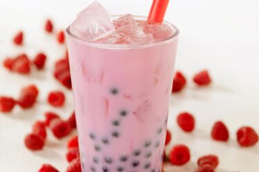 A tall glass of pink bubble tea with black tapioca pearls and ice cubes, served with a red straw. Fresh raspberries are scattered around the glass on a white surface, inspiring you to make bubble tea at home. The softly blurred background draws focus to the drink and fruit.