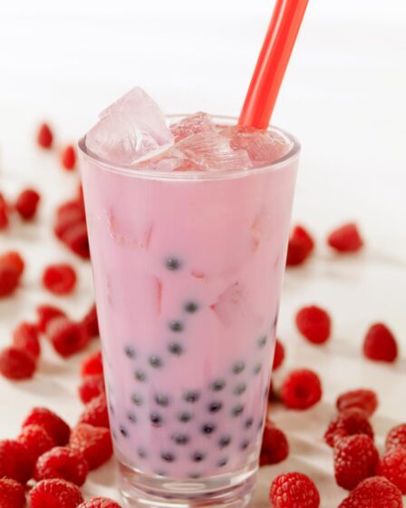 A tall glass of pink bubble tea with black tapioca pearls and ice cubes, served with a red straw. Fresh raspberries are scattered around the glass on a white surface, inspiring you to make bubble tea at home. The softly blurred background draws focus to the drink and fruit.