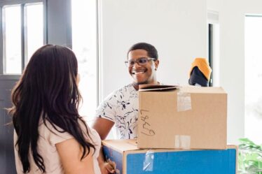A smiling man holds two cardboard boxes and a trainer by an open door, greeting a woman with long dark hair. Sunlight streams in, a green plant’s nearby—both look happy and relaxed, capturing the spirit of autumn house removal.