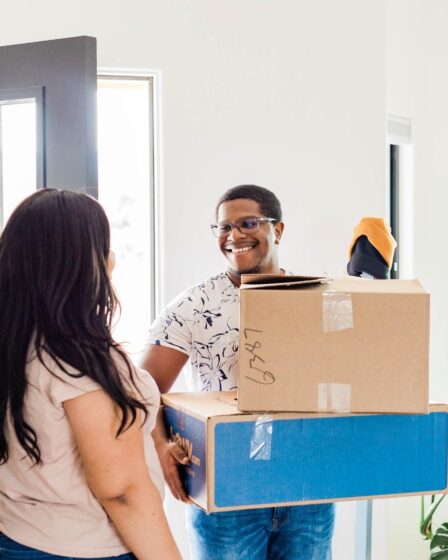 A smiling man holds two cardboard boxes and a trainer by an open door, greeting a woman with long dark hair. Sunlight streams in, a green plant’s nearby—both look happy and relaxed, capturing the spirit of autumn house removal.