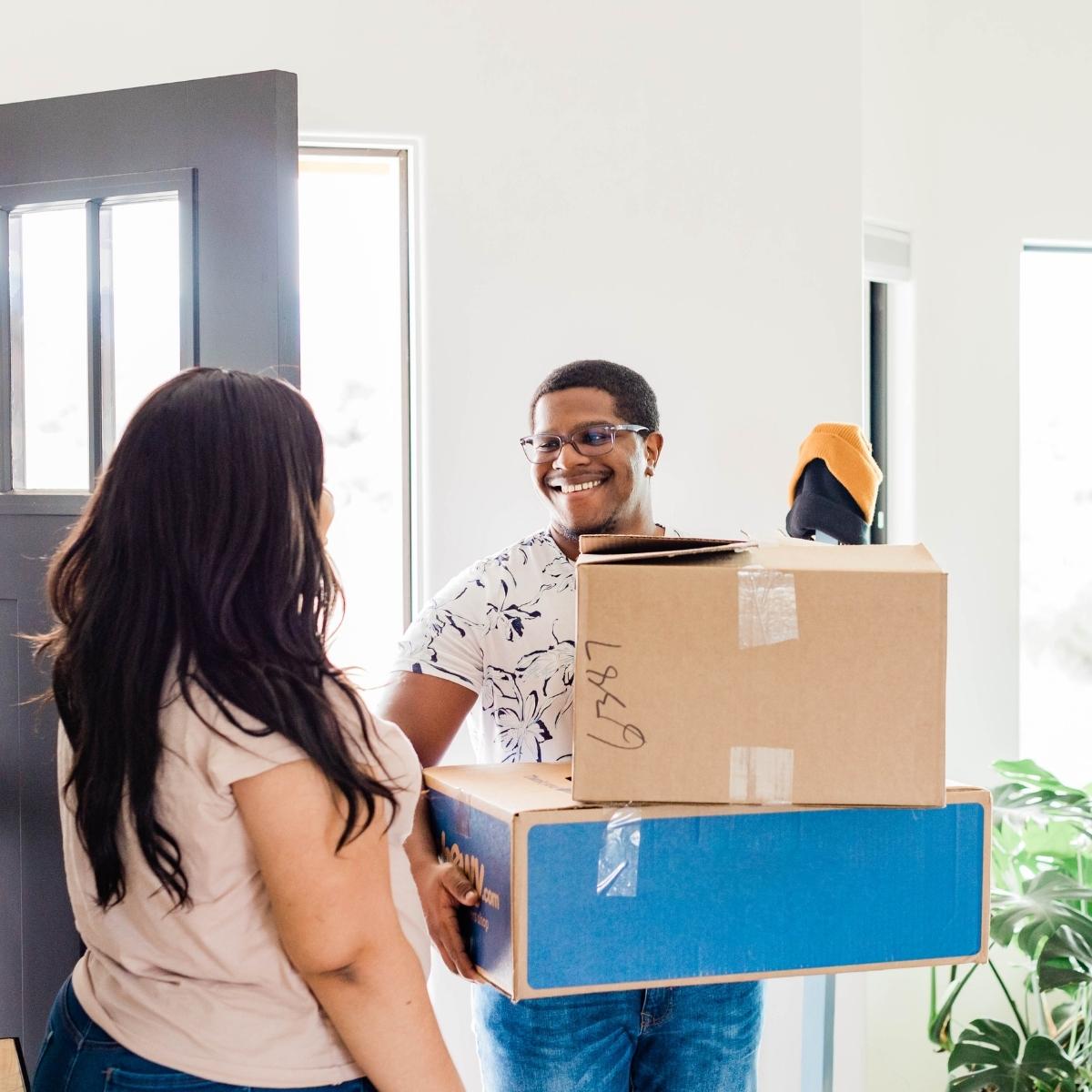 A smiling man holds two cardboard boxes and a trainer by an open door, greeting a woman with long dark hair. Sunlight streams in, a green plant’s nearby—both look happy and relaxed, capturing the spirit of autumn house removal.