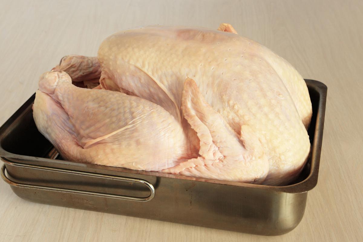 A whole raw turkey with pale, unseasoned skin sits in a large rectangular metal roasting tin on a light wood surface, ready for Christmas preparation—handle with care to avoid food poisoning.